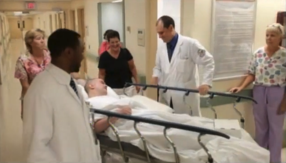 Hospital staff in white coats and scrubs walking alongside a wounded service member on a gurney down a VA medical center corridor, in a still from the VA documentary From Surviving to Thriving